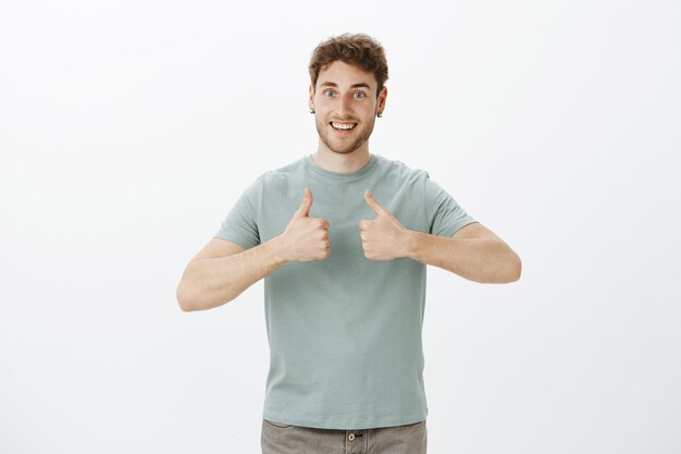 Porträt des aufgeregten glücklichen europäischen Kerls mit dem blonden Haar im T-Shirt, das Daumen hoch zeigt und breit lächelt, froh, ausgezeichnete Idee zu erhalten