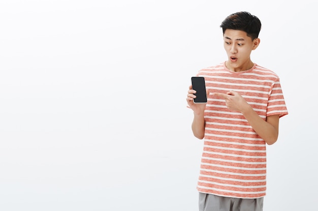 Porträt des beeindruckten und überraschten aufgeregten jungen attraktiven chinesischen Kerls im gestreiften T-Shirt offener Mund vom Interesse und vom Nervenkitzel, das Smartphone zeigt und auf Handybildschirm fasziniert hält