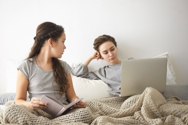 Porträt des brünetten Mädchens im grauen T-Shirt, das Buch im Bett liest und beobachtet, wie ihre junge Mutter im Hintergrund im Internet surft und ernsthaften Ausdruck hat. Menschen, Freizeit, Technologie und Kommunikation