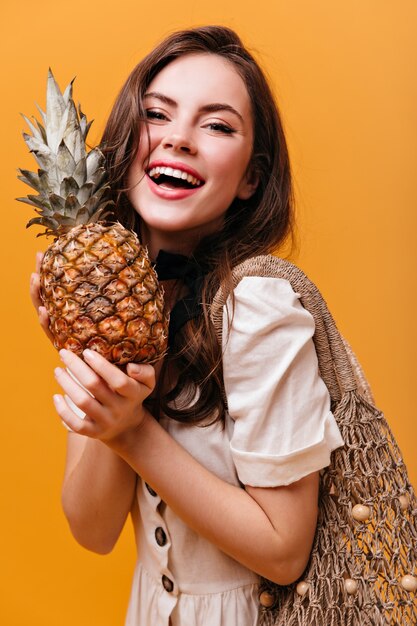 Porträt des brünetten Mädchens in ausgezeichneter Stimmung, die Ananas und Schnurbeutel auf orange Hintergrund hält.