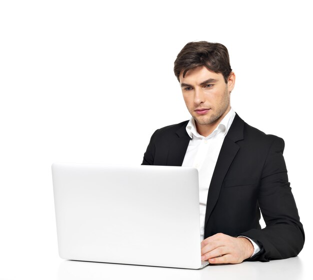 Porträt des denkenden jungen Büroangestellten mit Laptop, der auf Tisch lokalisiert auf Weiß sitzt.