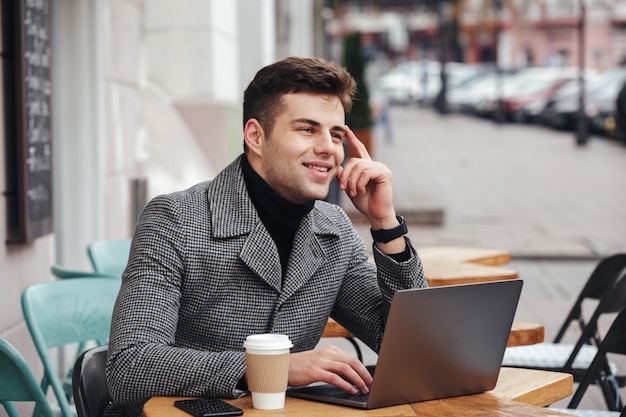 Porträt des erfolgreichen Kerls arbeitend mit silbernem Laptop im Straßencafé, an Geschäft denkend oder mit Freund plaudernd