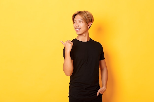 Porträt des erfreuten nachdenklichen asiatischen Kerls, der schaut und zeigt Finger links auf Logo, stehende gelbe Wand