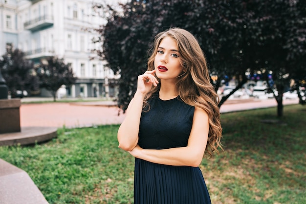 Porträt des ernsten eleganten Mädchens im Stadtpark. Sie hat lange Haare, ein schwarzes Kleid, weinige Lippen und sieht wehmütig aus.