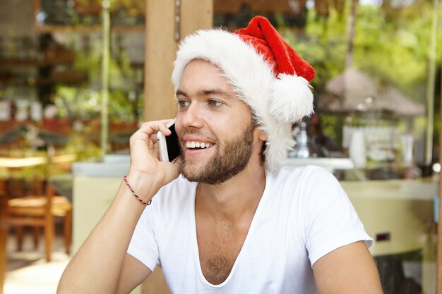 Porträt des freudigen hübschen jungen Mannes im weißen T-Shirt und im Weihnachtsmannhut, die auf Handy sprechen, während sie Ferien im tropischen Land genießen