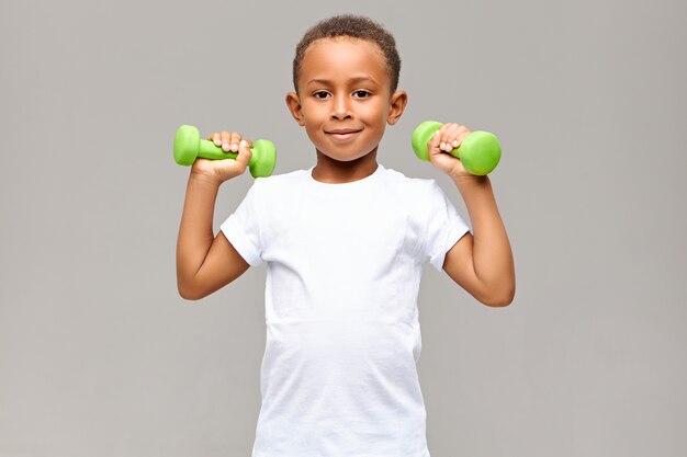 Porträt des fröhlichen afroamerikanischen Jungen mit den dünnen Armen, die glücklich lächeln, während sie im Fitnessstudio mit zwei Hanteln trainieren, um starken gesunden sportlichen Körper aufzubauen. Fitness und Kinder