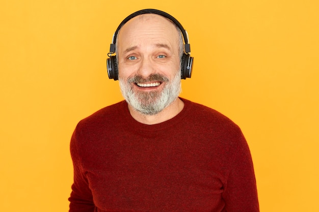 Porträt des fröhlichen emotionalen älteren Mannes mit dem dicken grauen Bart, der unter Verwendung der drahtlosen Kopfhörer lokalisiert aufwirft