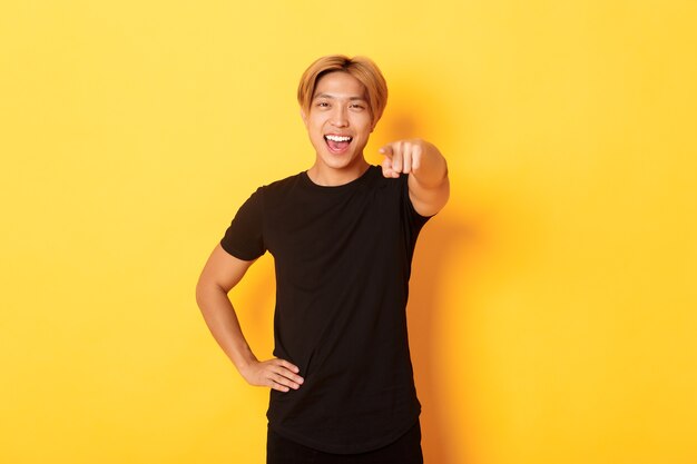 Porträt des fröhlichen gutaussehenden asiatischen Kerls mit blondem Haar, das Sie wählt, lächelnd und Finger zeigt, Glückwunschgeste.