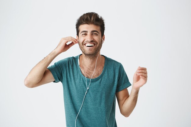 Porträt des fröhlichen hispanischen Kerls im blauen T-Shirt, das Kopfhörer mit Hand hält, Musik hört und mit glücklichem Gesichtsausdruck tanzt. Positive Emotionen und Humor.