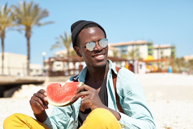 Porträt des fröhlichen jungen Mannes, der sich am städtischen Strand entspannt, der Scheibe der reifen Wassermelone hält
