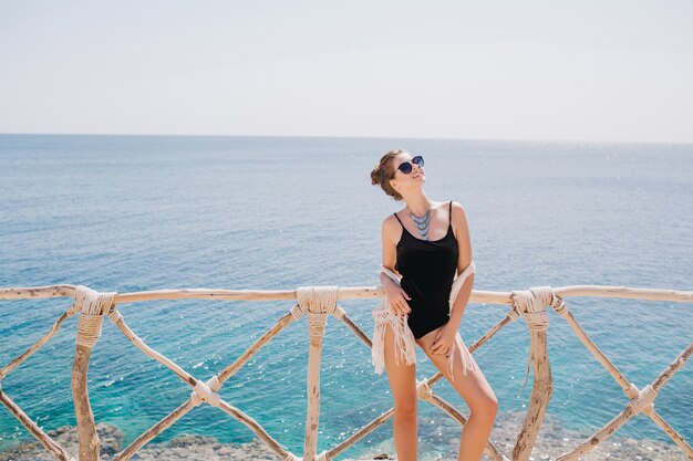 Porträt des fröhlichen Mädchens in der Sonnenbrille, die Sonne und frische Luft auf dem Seebad im Sommertag genießt. Erstaunliche junge Frau mit niedlicher Frisur im schwarzen Badeanzug, der nahe dem Ozean aufwirft
