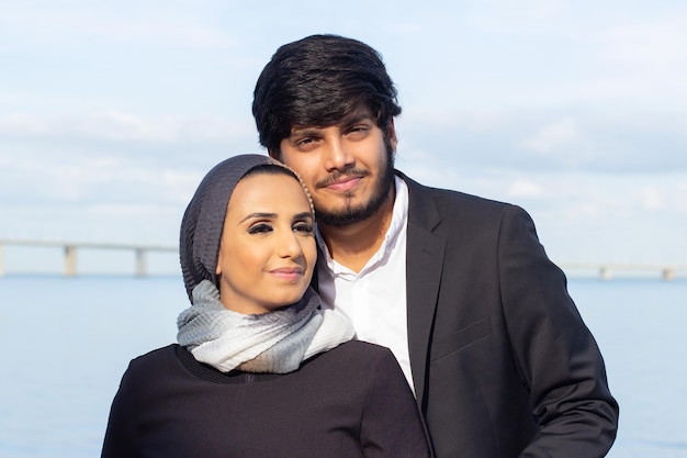 Porträt des fürsorglichen arabischen Paares während des Spaziergangs. Frau mit bedecktem Kopf und hellem Make-up und Mann im Anzug, der in die Kamera lächelt und sich umarmt. Liebe, Zuneigung Konzept
