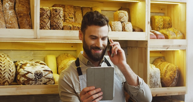 Porträt des glücklichen bäckers, der im bäckereigeschäft unter verwendung des digitalen tablet-computers arbeitet und auf handy spricht