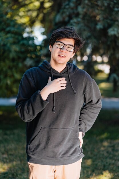 Porträt des glücklichen jungen männlichen Studenten mit Brille im lässigen Outfit, das am Park aufwirft.