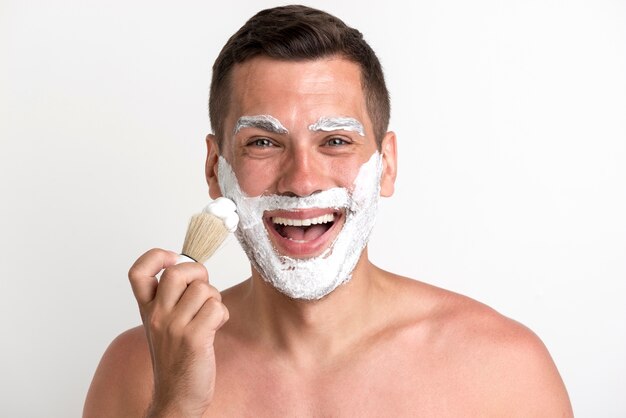 Porträt des glücklichen jungen Mannes, der Schaum gegen weißen Hintergrund rasierend zutrifft