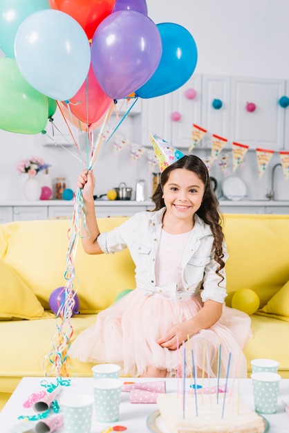 Kostenloses Foto porträt des glücklichen mädchens sitzend auf dem sofa, das bunte ballone hält