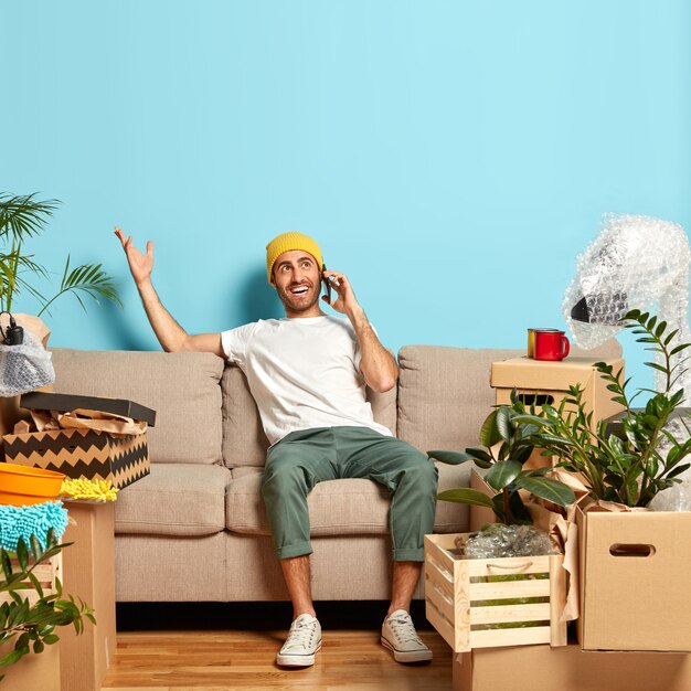 Porträt des glücklichen Mannes hat Telefongespräch, Gesten mit einer Hand, versucht den Weg zu seiner neuen Wohnung zu erklären, trägt gelben Hut