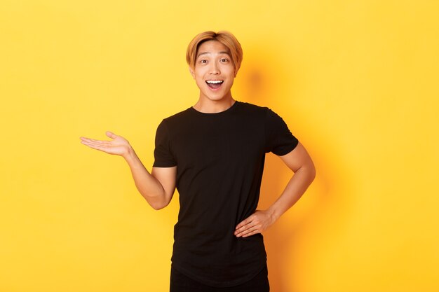 Porträt des glücklichen und aufgeregten lächelnden asiatischen Kerls, der etwas auf der Hand über gelber Wand hält