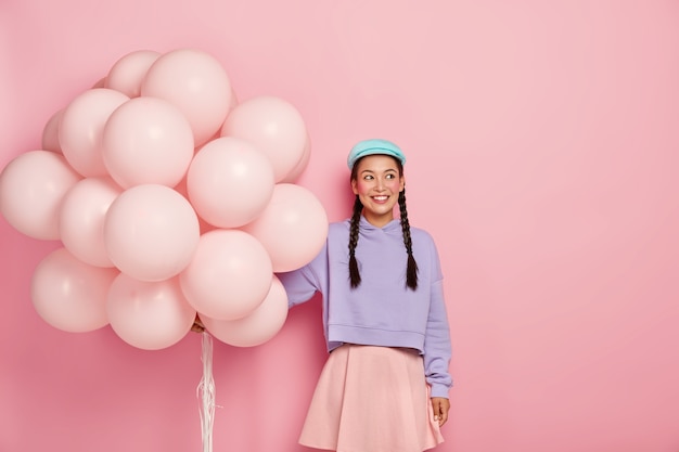 Porträt des glücklichen zufriedenen Mädchens mit langen Zöpfen, trägt losen Pullover, Rock, hat minimales Make-up, steht mit aufgeblasenen Luftballons gegen rosa Wand