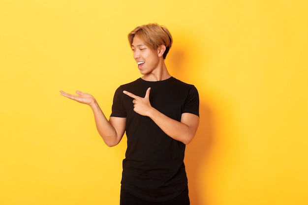 Porträt des gutaussehenden lächelnden asiatischen Kerls, der etwas auf der Hand hält und Finger darauf zeigt, gelbe Wand erfreut erfreut.