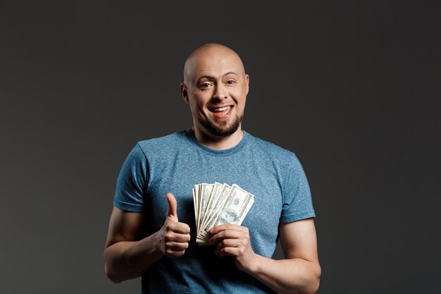 Porträt des gutaussehenden Mannes im grauen Hemd, das Geld über dunkler Wand hält