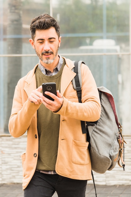 Porträt des gutaussehenden Mannes mit seinem Rucksack unter Verwendung des Smartphone