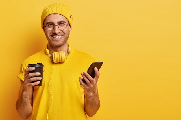 Porträt des hübschen fröhlichen jungen Mannes mit erfreutem Gesichtsausdruck, hält Handy, sendet Textnachrichten an Freunde, trinkt Kaffee zum Mitnehmen, trägt eine Brille, gelbes Outfit mit Kopfhörern