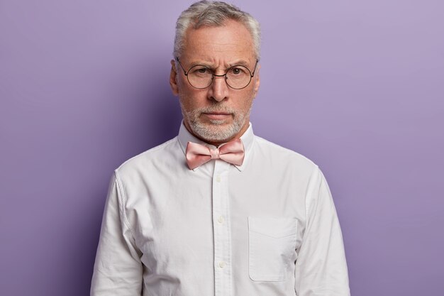 Porträt des hübschen grauhaarigen europäischen Mannes sieht mit striktem Gesichtsausdruck durch runde Brille aus, trägt formelles weißes Hemd und Fliege