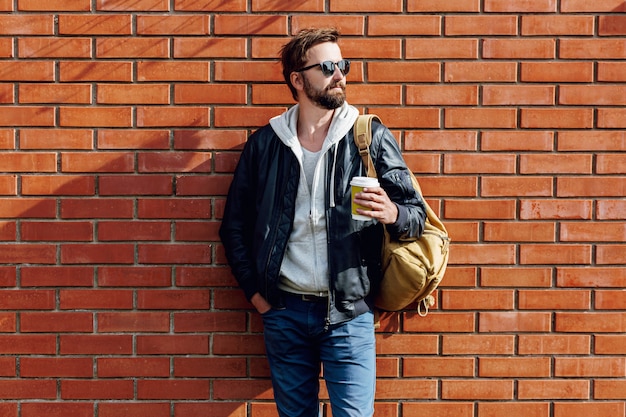 Porträt des hübschen Hipster-Mannes mit Bart im Hut, grauem Kapuzenpulli und Lederjacke mit Beutelpackung, die Tasse heißen Kaffee hält