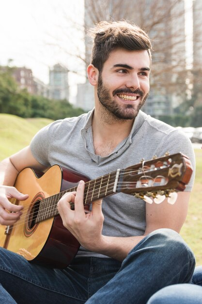 Porträt des hübschen jungen Mannes, der Gitarre im Park spielt