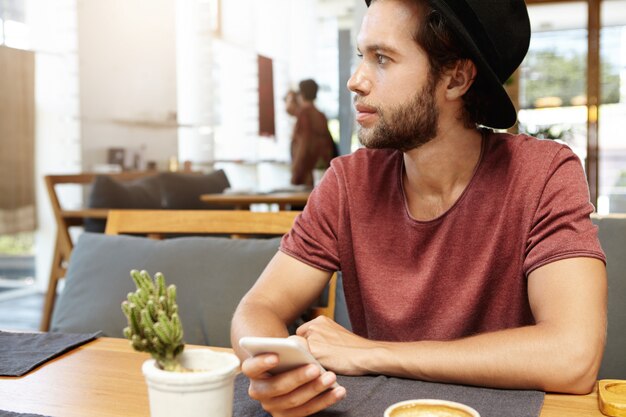 Porträt des hübschen jungen Mannes mit Stoppeln, die am Holztisch sitzen und generisches Smartphone halten