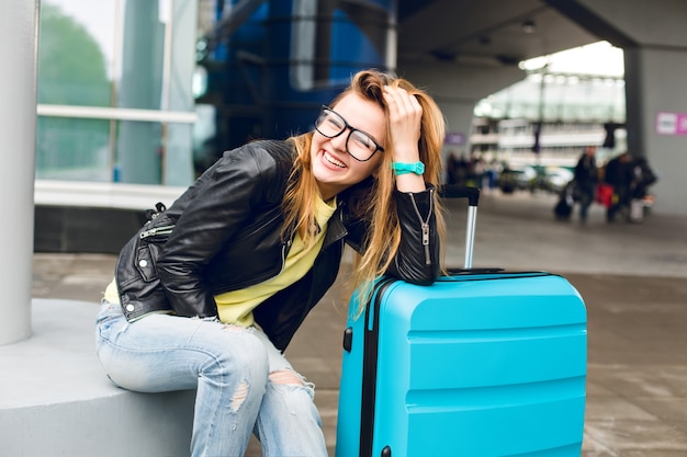 Porträt des hübschen Mädchens mit den langen Haaren in den Gläsern, die draußen im Flughafen sitzen. Sie trägt einen gelben Pullover mit schwarzer Jacke und Jeans. Sie beugte sich zum Koffer und lächelte in die Kamera.