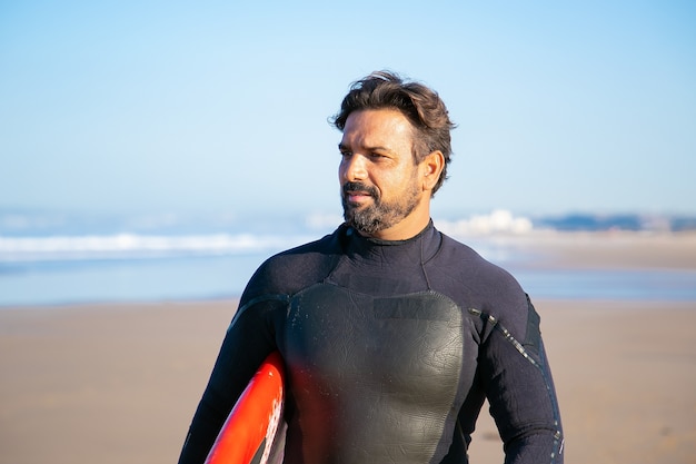 Porträt des hübschen Surfers, der am Strand mit Surfbrett steht und wegschaut