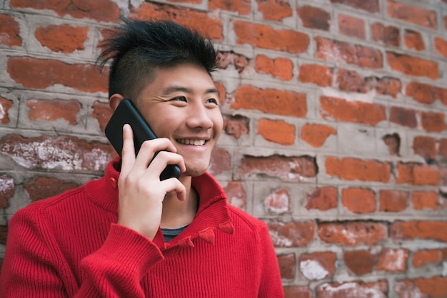 Porträt des jungen asiatischen Mannes, der am Telefon draußen gegen Backsteinmauer spricht. Kommunikationskonzept.