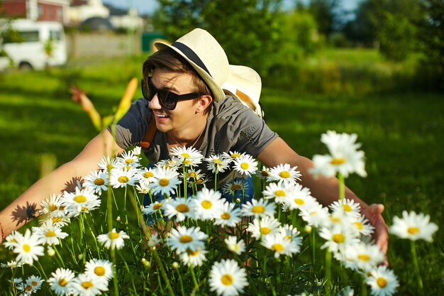 Porträt des jungen attraktiven lächelnden modernen stilvollen Mannes im zufälligen Stoff im Hut in den Gläsern im Park mit hellen bunten Blumen in der Kamille