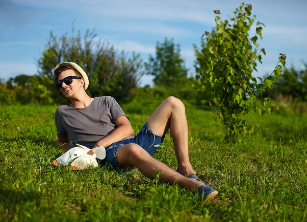 Porträt des jungen attraktiven modernen stilvollen Mannes im lässigen Stoff im Hut in den Gläsern, die im Park im grünen Gras sitzen