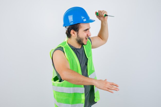 Porträt des jungen Baumeisters, der Hand hebt, um mit Schraubendreher in Uniform zu schlagen und verrückte Vorderansicht zu schauen