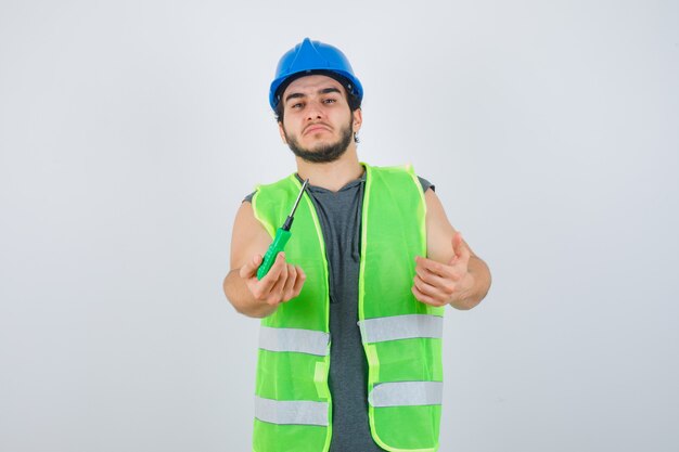 Porträt des jungen Baumeisters, der Hand streckt, um Schraubendreher in Uniform zu geben und selbstbewusste Vorderansicht zu schauen