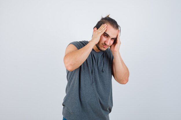 Porträt des jungen fitten Mannes, der unter Kopfschmerzen im ärmellosen Kapuzenpulli leidet und schmerzhafte Vorderansicht schaut