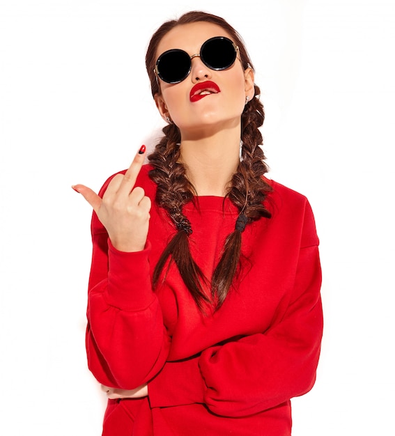 Porträt des jungen glücklichen lächelnden Frauenmodells mit hellem Make-up und den bunten Lippen mit zwei Zöpfen und Sonnenbrille in der roten Kleidung des Sommers lokalisiert. Fuck-Off-Zeichen zeigen