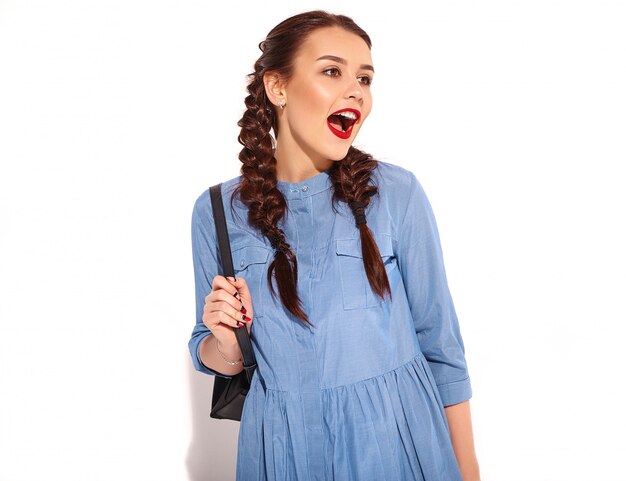Porträt des jungen glücklichen lächelnden Frauenmodells mit hellem Make-up und den roten Lippen mit zwei Zöpfen in den Händen im bunten blauen Kleid und im Rucksack des Sommers lokalisiert