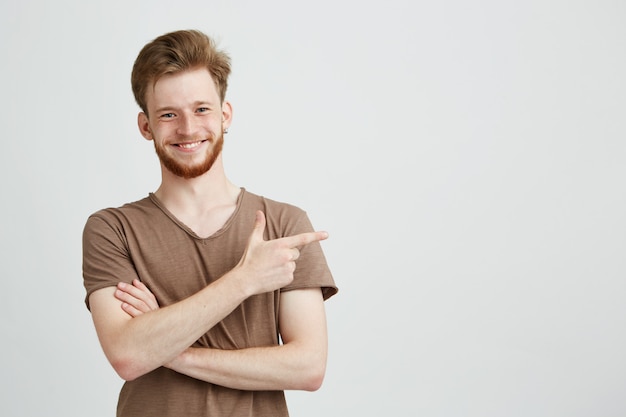 Porträt des jungen gutaussehenden aufrichtigen mannes mit dem lächelnden bart, der finger in seite zeigt.