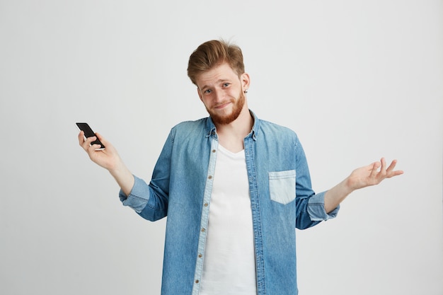 Porträt des jungen gutaussehenden Kerls, der Smartphone-Achselzucken hält und Kamera betrachtet.