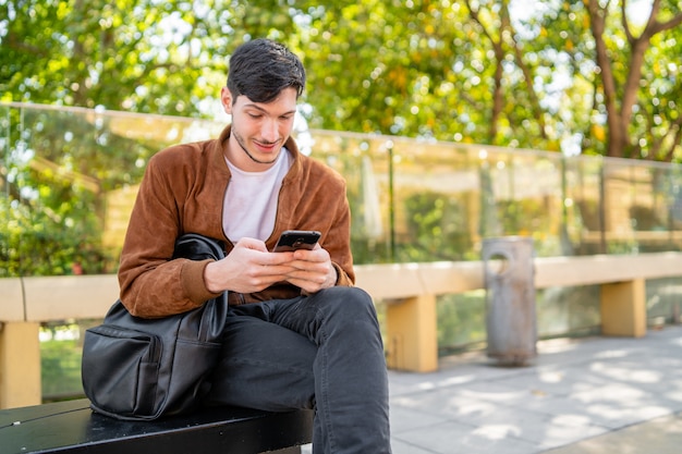 Porträt des jungen gutaussehenden Mannes, der sein Handy benutzt, während er draußen sitzt. Kommunikation und Stadtkonzept.