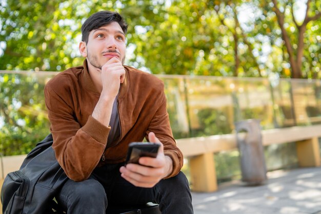 Porträt des jungen gutaussehenden Mannes, der sein Handy benutzt, während er draußen sitzt. Kommunikation und Stadtkonzept.