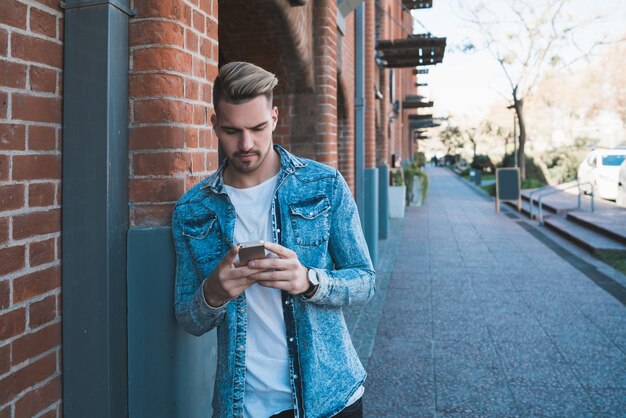 Porträt des jungen gutaussehenden Mannes, der sein Handy draußen in der Straße benutzt. Kommunikationskonzept.