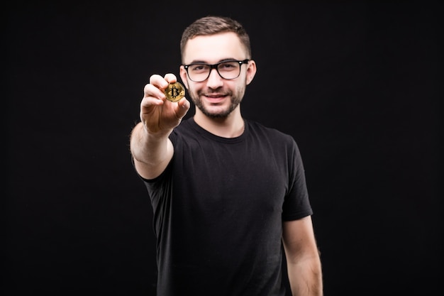 Porträt des jungen gutaussehenden Mannes in den Gläsern im schwarzen Hemd zeigte goldenes Bitcoin auf Kamera lokalisiert auf Schwarz