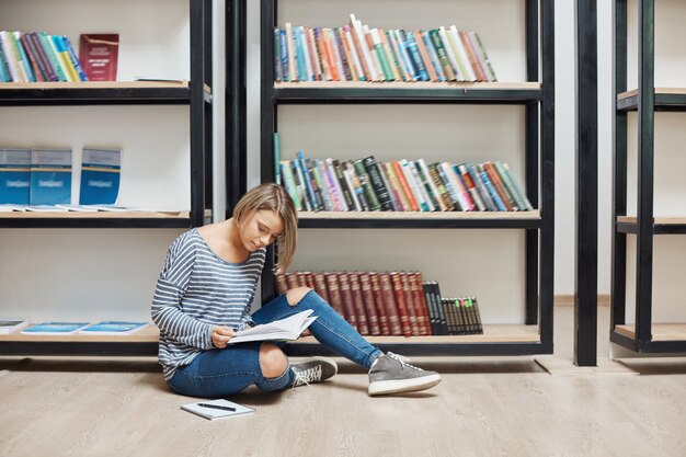 Porträt des jungen gutaussehenden Studentenmädchens mit kurzen blonden Haaren in lässiger, stilvoller Kleidung, die auf dem Boden in einer modernen Bibliothek in der Nähe von Regalen sitzt, Lieblingsbücher liest und ein Wochenende in gemütlicher Atmosphäre verbringt