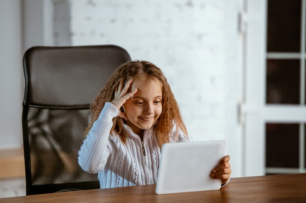 Porträt des jungen kaukasischen Mädchens sieht verträumt, niedlich und glücklich aus. Nach oben schauen, drinnen am Holztisch mit Tablet und Smartphone sitzen. Konzept der Zukunft, Ziel, Traum zu kaufen, Visualisierung.