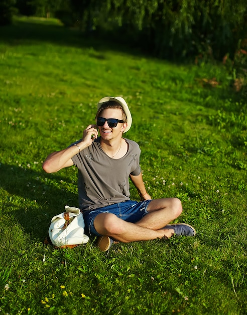 Porträt des jungen lächelnden lachenden attraktiven modernen stilvollen Mannes im lässigen Stoff im Hut in den Gläsern, die im Park im grünen Gras sitzen am Telefon sprechen